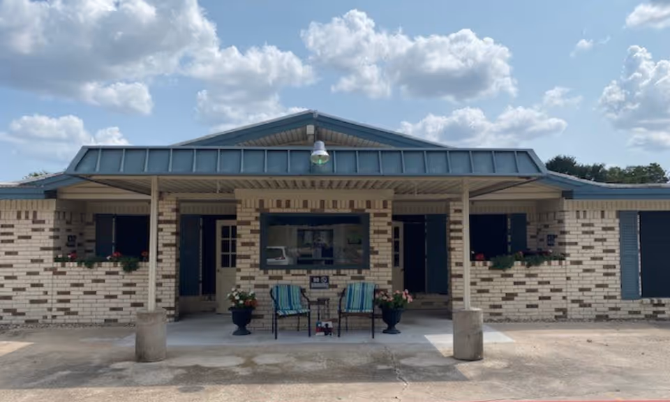 Single-story brick senior living facility front entrance with a covered porch, two chairs, potted plants, and a blue metal roof under a partly cloudy sky.