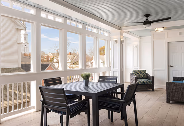 A bright and airy enclosed porch area with large windows showing a view of nearby houses and a church steeple. The space features a black dining table with four matching chairs, a potted plant centerpiece, two cushioned armchairs with patterned pillows, a ceiling fan, and light wood flooring.
