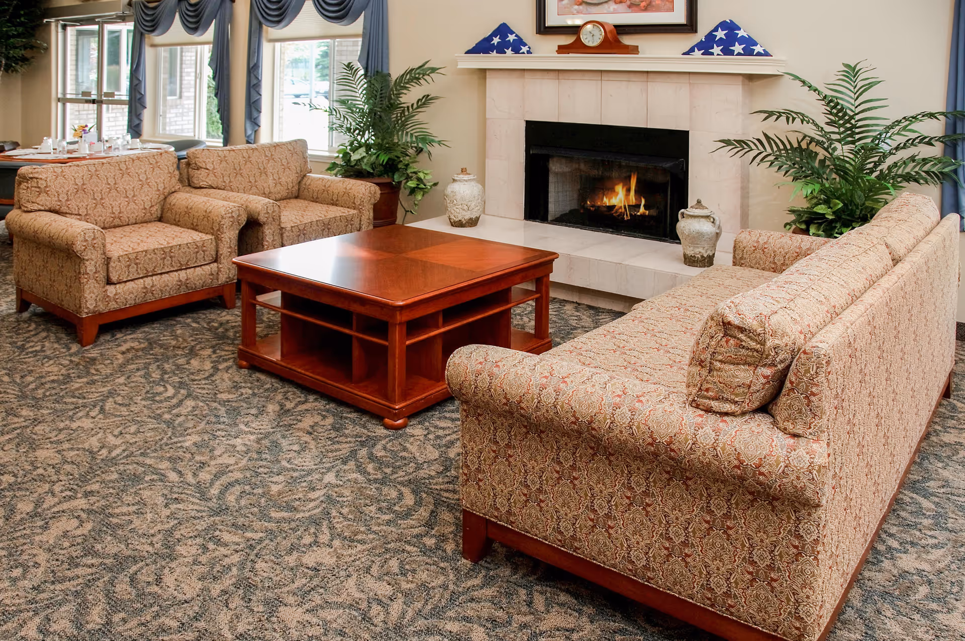 A cozy living room area with a patterned sofa and two matching armchairs arranged around a wooden coffee table. There is a lit fireplace with a mantel above it, decorated with a clock and folded American flag displays. Large windows with blue curtains let in natural light, and potted plants add greenery to the space.