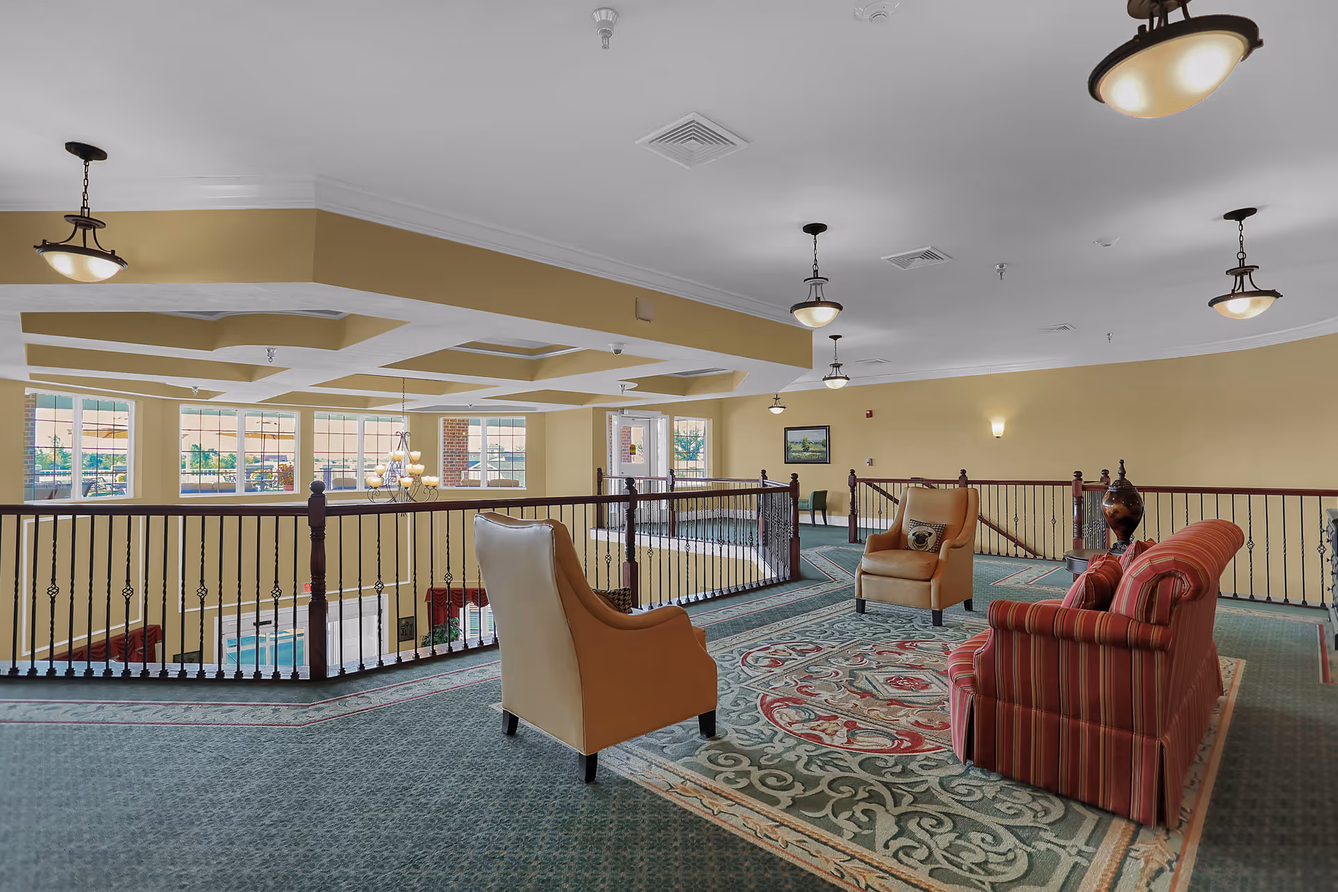 A spacious indoor common area with carpeted floors and a decorative rug. The area features comfortable seating including two armchairs and a striped sofa arranged around a small table. The space overlooks a lower level through a railing, with large windows allowing natural light to fill the room. Ceiling lights and wall sconces provide additional illumination.