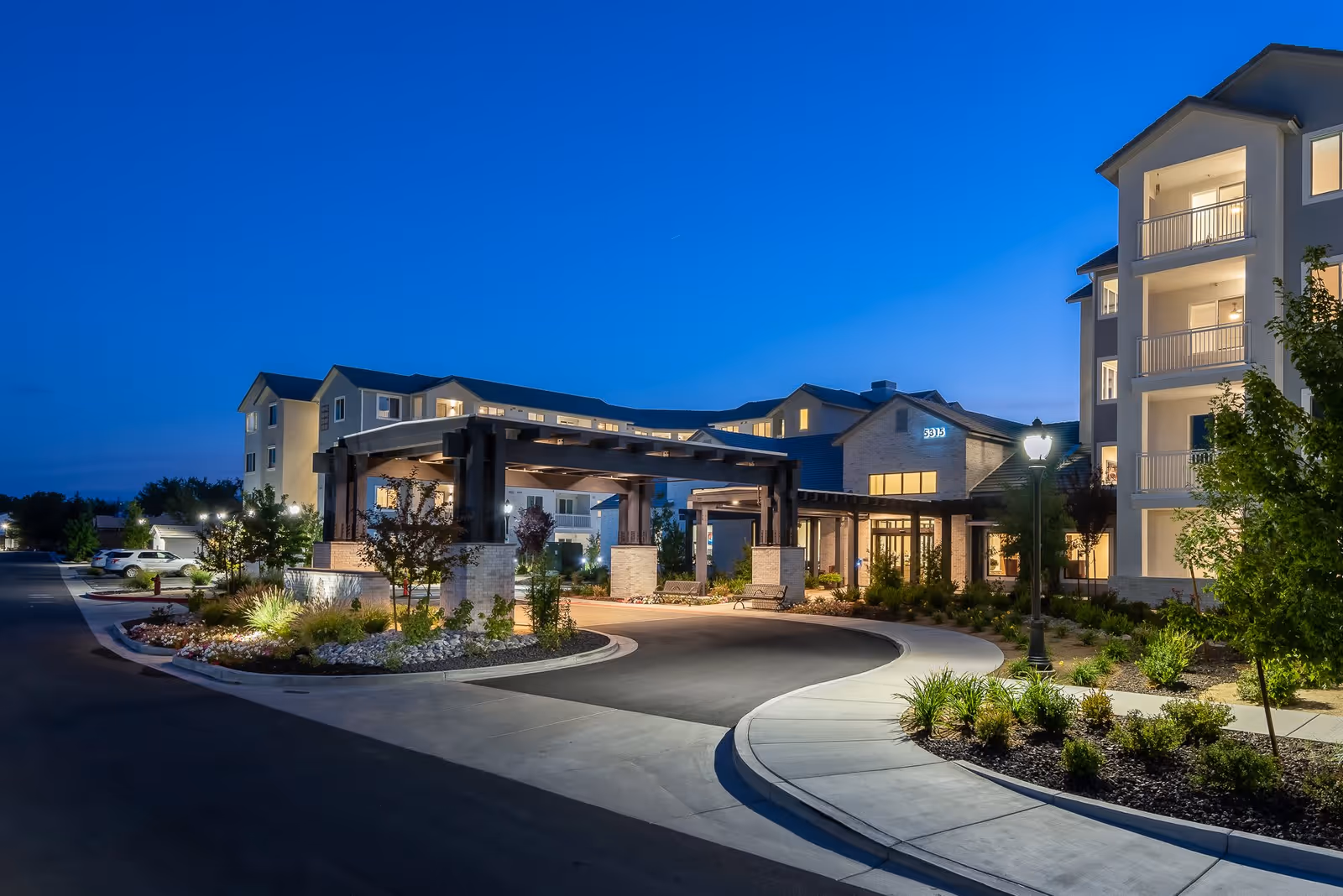 Exterior view of a senior living facility named Revel Rancharrah at dusk, showing a well-lit entrance with a covered driveway, landscaped garden areas, and multi-story buildings with balconies.