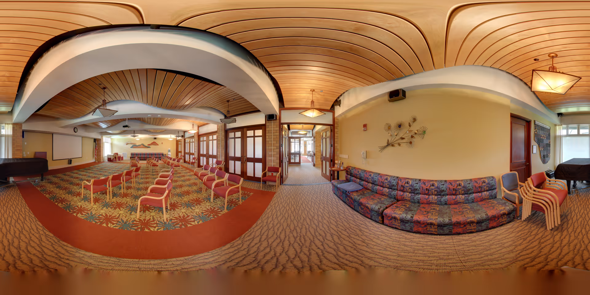 Panoramic view of a senior living facility common area with rows of red chairs arranged facing a stage with a podium and a large screen. The room features a patterned carpet, a curved wooden ceiling, and a long multicolored couch along one wall. There are decorative wall hangings and multiple doors leading to other areas.