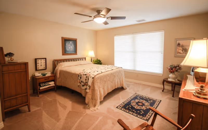 A cozy bedroom with a bed covered in a beige quilt and a patterned throw blanket. The room has beige walls and carpet, a ceiling fan with lights, a large window with blinds, a wooden dresser, a nightstand with a lamp and a phone, a small side table with a vase of flowers, and framed artwork on the walls.