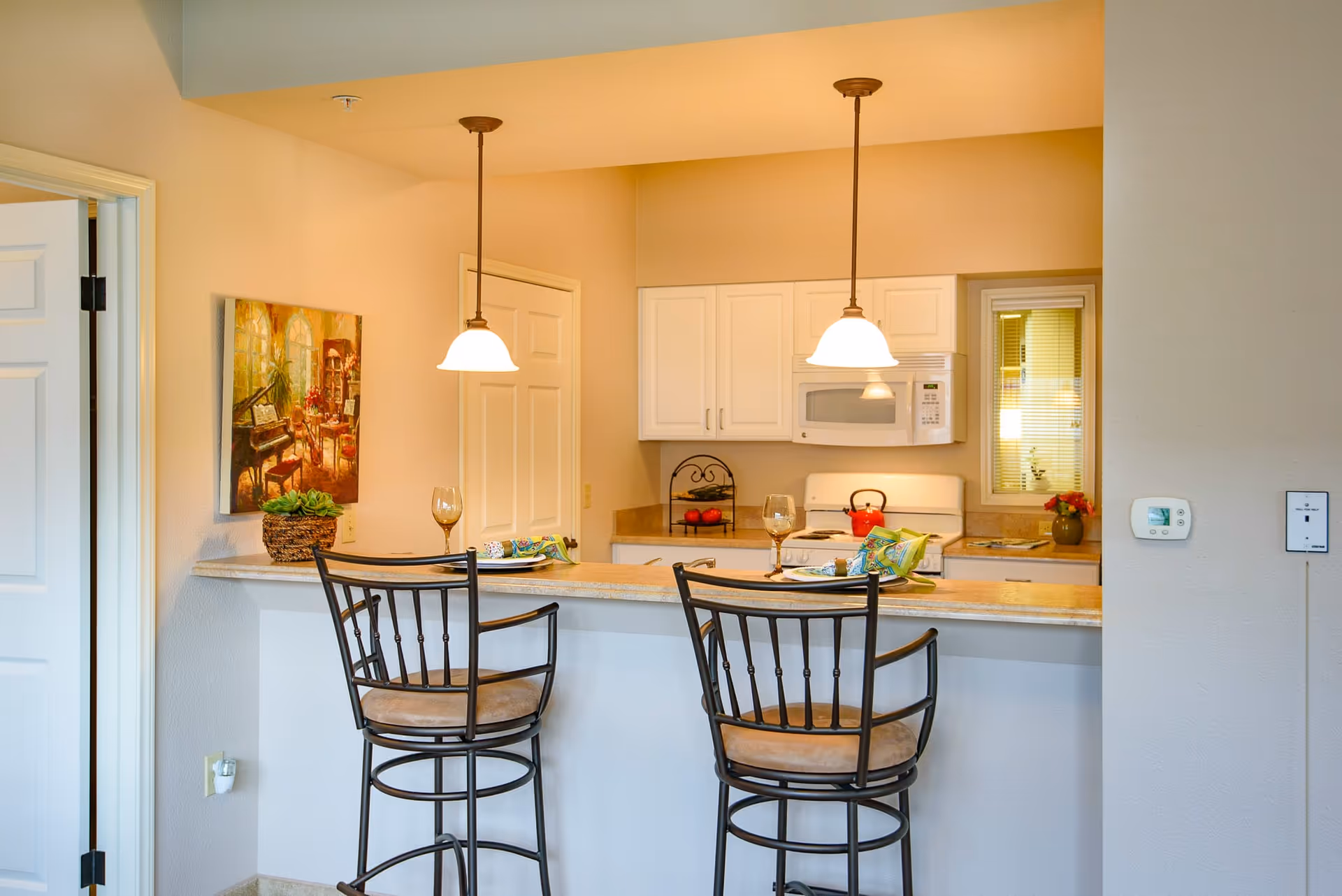 A cozy kitchen area with a breakfast bar featuring two high chairs with cushioned seats. The kitchen has white cabinets, a microwave, and a stove with a red kettle on it. Two pendant lights hang above the bar, which is set with plates, napkins, and wine glasses. A painting and a small plant are on the left wall, and a thermostat is mounted on the right wall.