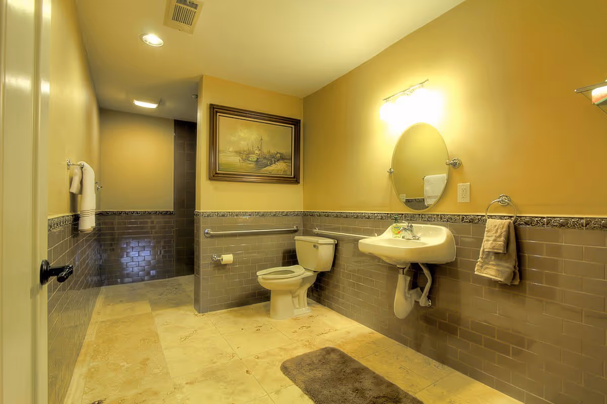 A spacious bathroom with beige walls and tiled lower half in gray. The room features a white toilet, a wall-mounted sink with a round mirror above it, and a towel ring with a beige towel. There is a framed painting on the wall above the toilet. The floor is tiled in a light beige color with a brown bath mat in front of the sink. The bathroom has recessed ceiling lights and a visible ventilation vent.