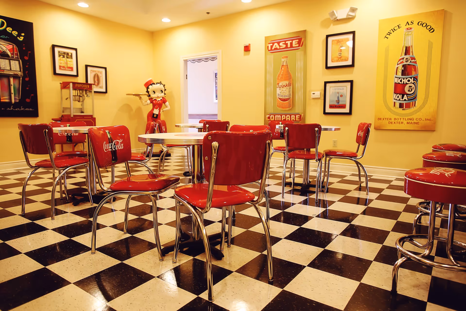 A retro-style dining area with black and white checkered floor, red Coca-Cola branded chairs, round tables, and vintage posters on yellow walls. A Betty Boop statue holding a tray and a popcorn machine are visible in the background.