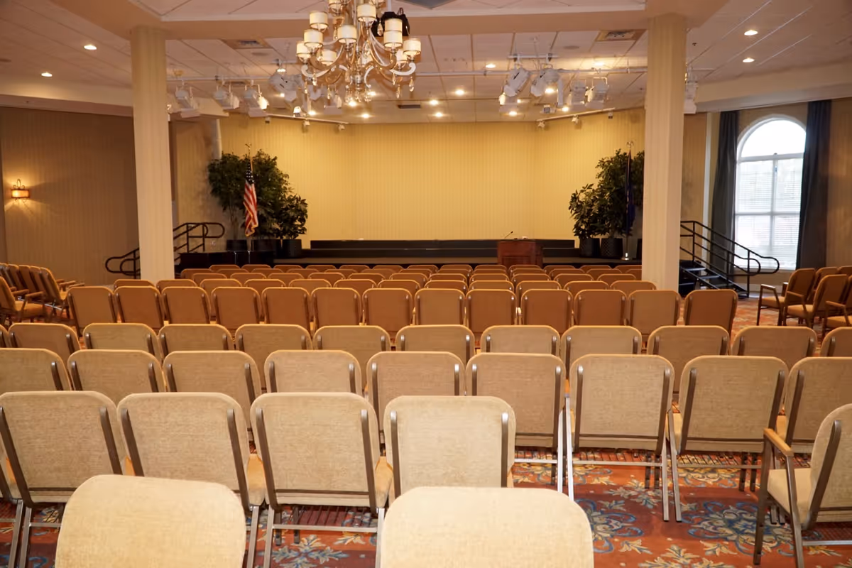 A large meeting or event room with rows of beige chairs facing a small stage with a podium. The room has beige walls, two large columns, chandeliers, and large windows with curtains. There are potted plants and an American flag on the stage.
