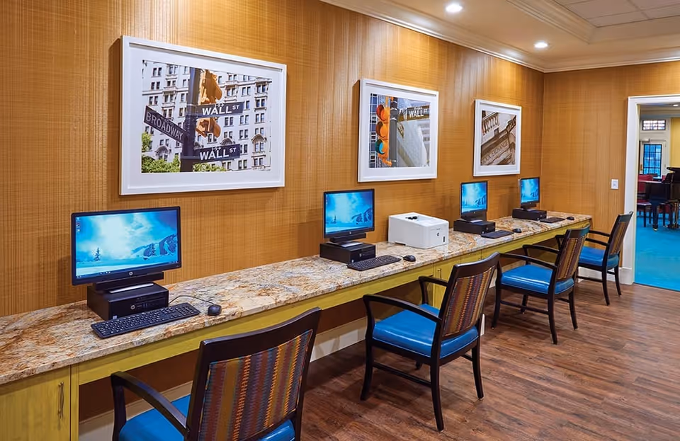 A computer room with four desktop computers and a printer on a long marble countertop. Four chairs with blue cushions are placed in front of the computers. The walls are covered with a textured brown wallpaper and decorated with framed pictures of street signs and traffic lights. The floor is wooden, and there is a doorway leading to another room with blue carpet.