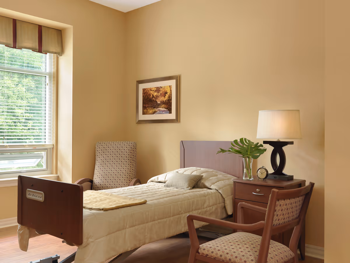 A cozy bedroom in a senior living facility with a single bed made up with beige bedding, a wooden headboard and footboard, a bedside table with a lamp, a small clock, and a vase with green leaves. There are two upholstered chairs with patterned fabric, a window with blinds and a valance, and a framed picture of a nature scene on the beige wall.