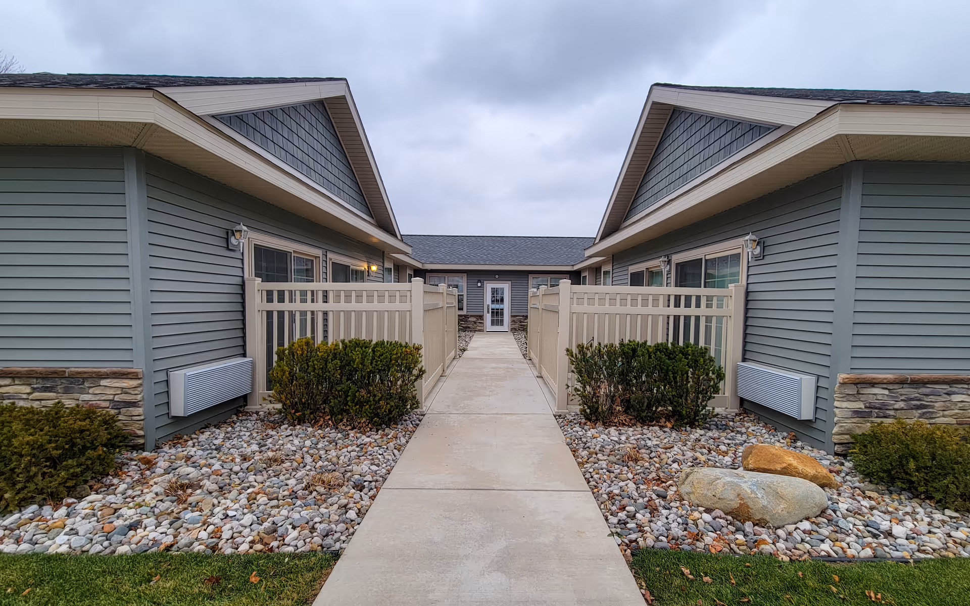 Exterior view of a senior living facility showing a paved walkway between two single-story buildings with gray siding and stone accents. There are small fenced patios with bushes and decorative rocks on either side of the walkway, leading to a door at the end. The sky is overcast.