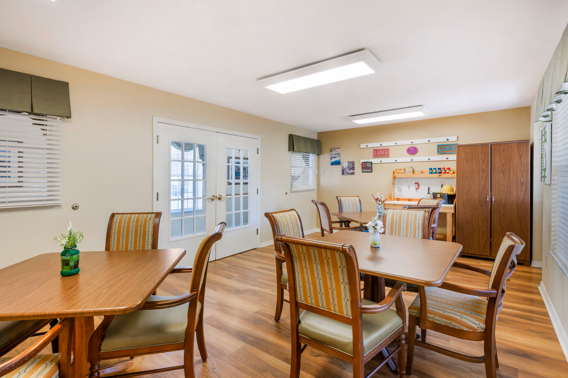Well-lit memory care dining/activity room with wooden tables and upholstered chairs, cabinets, and double French doors.