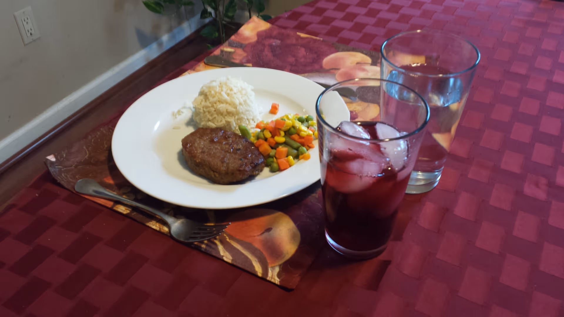 Plate with rice, mixed vegetables and a meat patty on a placemat next to two glasses (one with an iced red drink) on a red checkered tablecloth.