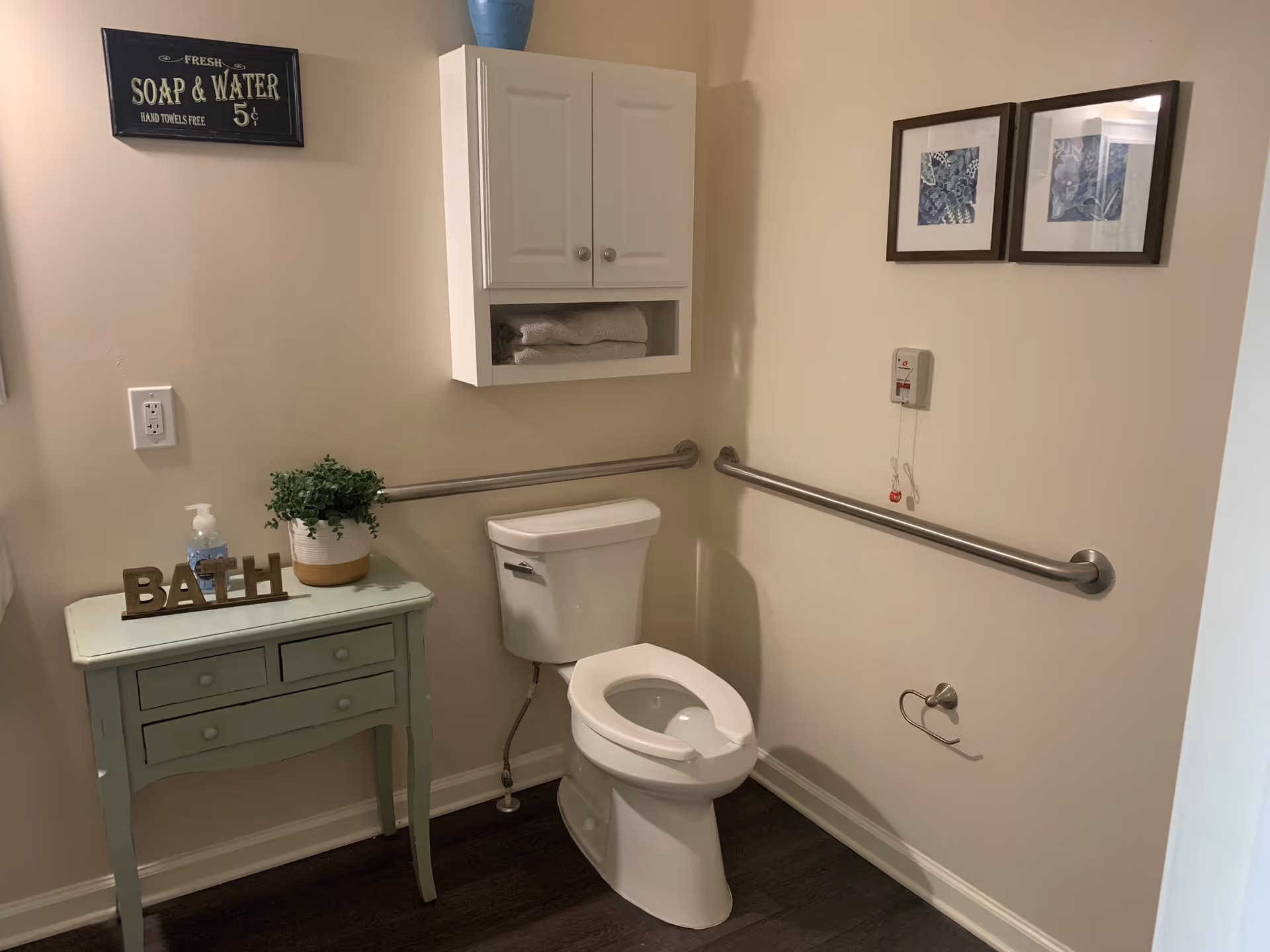 A clean and accessible bathroom with a white toilet, grab bars on two walls, a small green table with a plant, soap dispenser, and decorative letters spelling BATH. Above the toilet is a white cabinet with towels and two framed blue floral pictures on the wall.