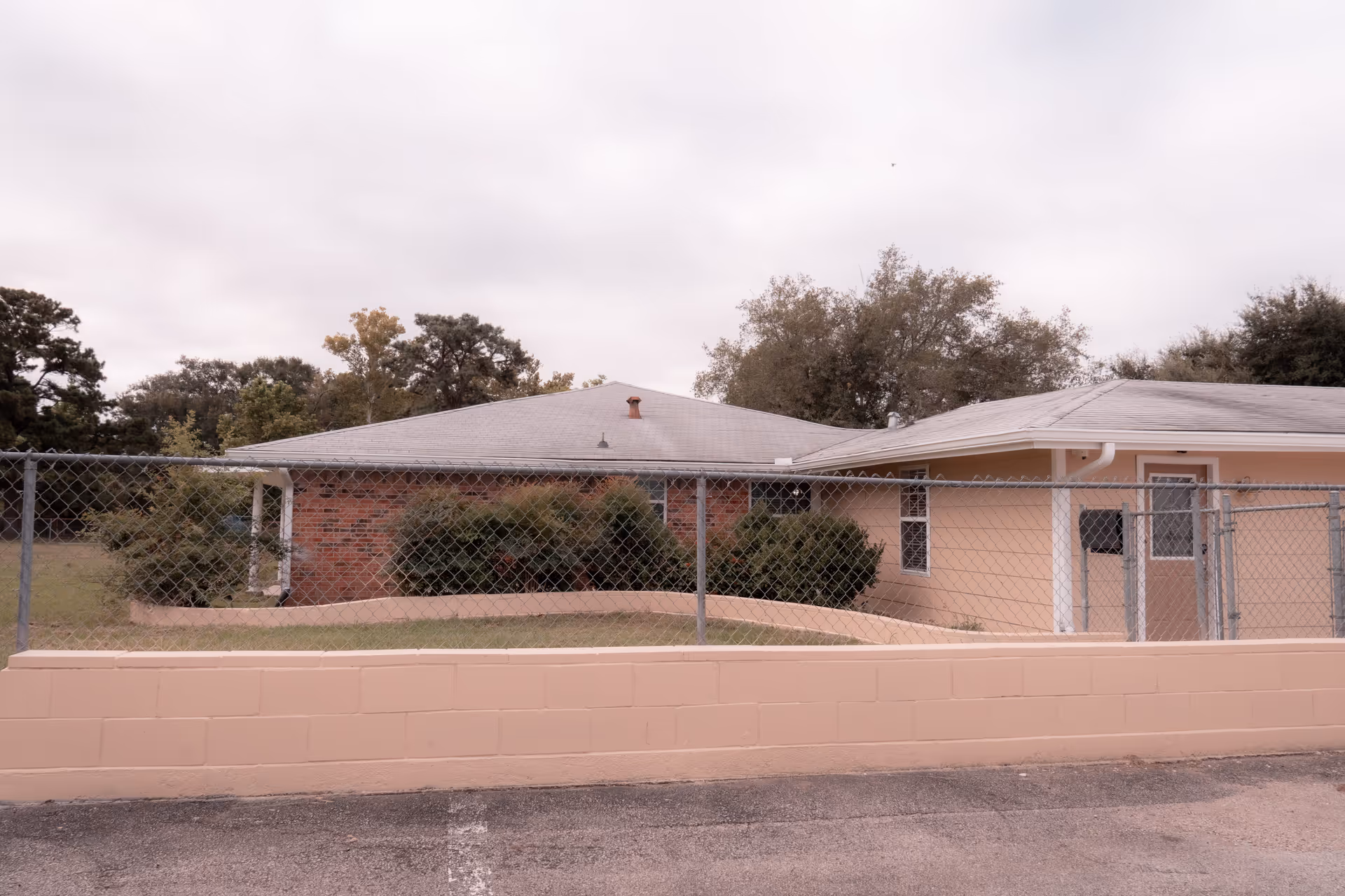 Single-story brick and stucco building behind a chain-link fence and low cinderblock wall under a cloudy sky.