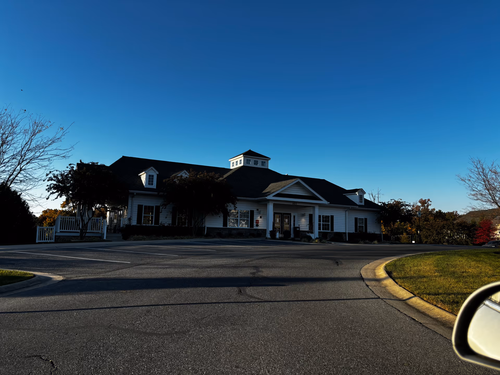 Exterior view of a single-story building with white siding and a dark roof under a clear blue sky. The building has a small cupola on the roof and several windows with black shutters. There is a paved driveway and parking area in front, with some trees and grass on the sides. The photo is taken from inside a vehicle, with a side mirror visible in the lower right corner.