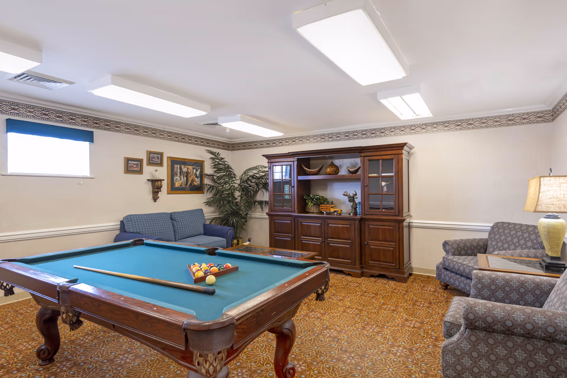 A recreational room with a pool table in the center, a blue couch against the wall, two patterned armchairs, a wooden cabinet with decorative items, a coffee table, a potted plant, and framed artwork on the walls. The room has patterned carpet and fluorescent ceiling lights.