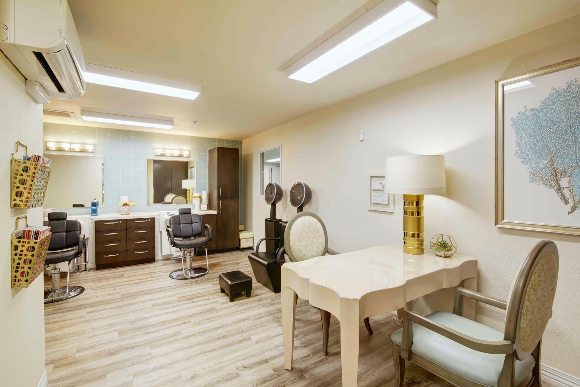 A bright and clean salon room in a senior living facility featuring two black salon chairs in front of mirrors with a cabinet and drawers, two hair dryers on stands, a white table with a gold lamp and two upholstered chairs, light wood flooring, and soft yellow walls with a framed artwork.