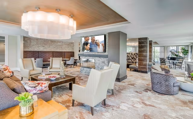 Bright, spacious communal lounge with grouped seating, a central fireplace with TV, chandelier, and a piano in the background.