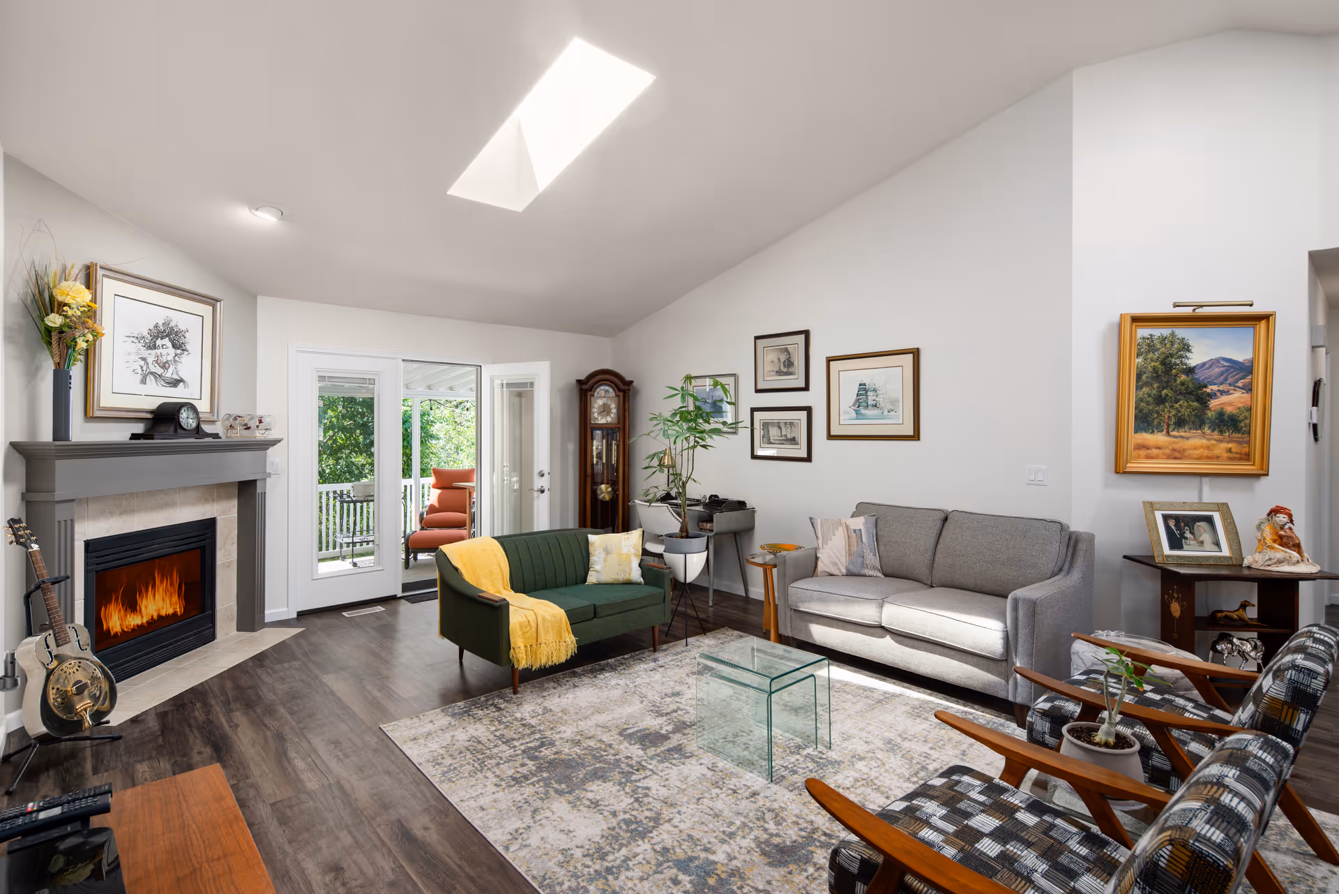 A bright and cozy living room with a green loveseat draped with a yellow throw blanket, a gray sofa, and two patterned armchairs. The room features a fireplace with a guitar leaning against it, framed artwork on the walls, a glass coffee table on a patterned rug, and a grandfather clock near glass doors leading to an outdoor patio with seating. There is a skylight in the ceiling allowing natural light to fill the space.