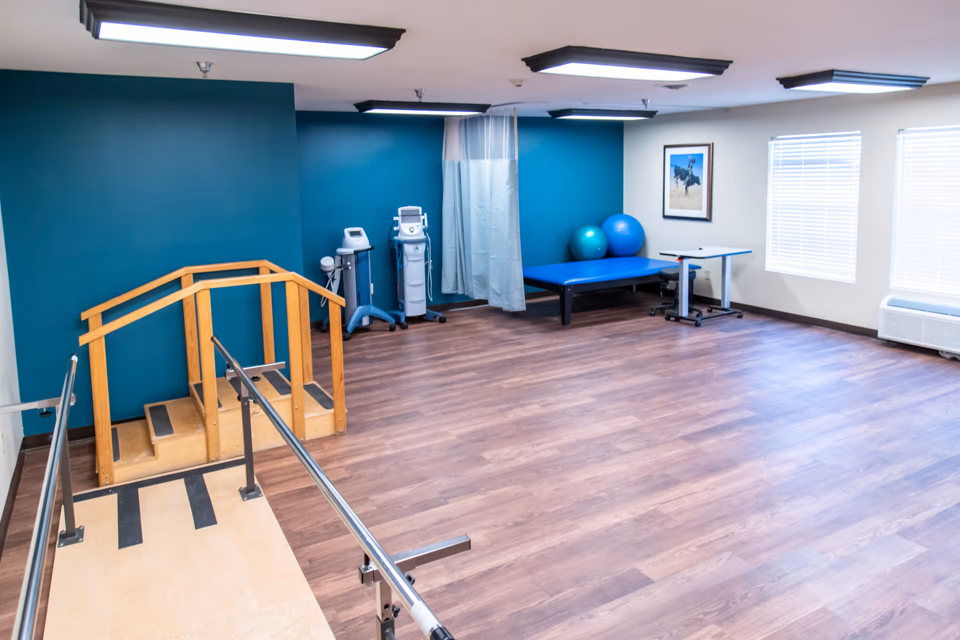A spacious rehabilitation room with wooden flooring, teal accent wall, and white walls with windows. The room contains parallel bars for walking exercises, a small wooden staircase, two exercise balls, a blue therapy table, medical equipment, and a small adjustable table on wheels. The room is well-lit with ceiling lights.