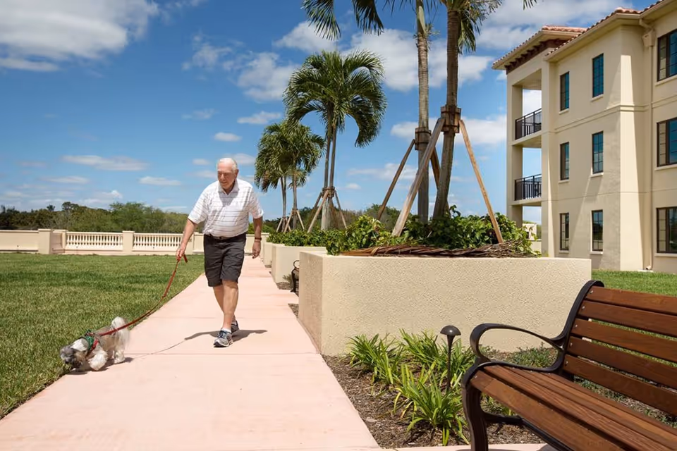 An elderly man walking a small dog on a leash along a paved pathway in a landscaped outdoor area with palm trees and a beige multi-story building in the background under a blue sky with scattered clouds.