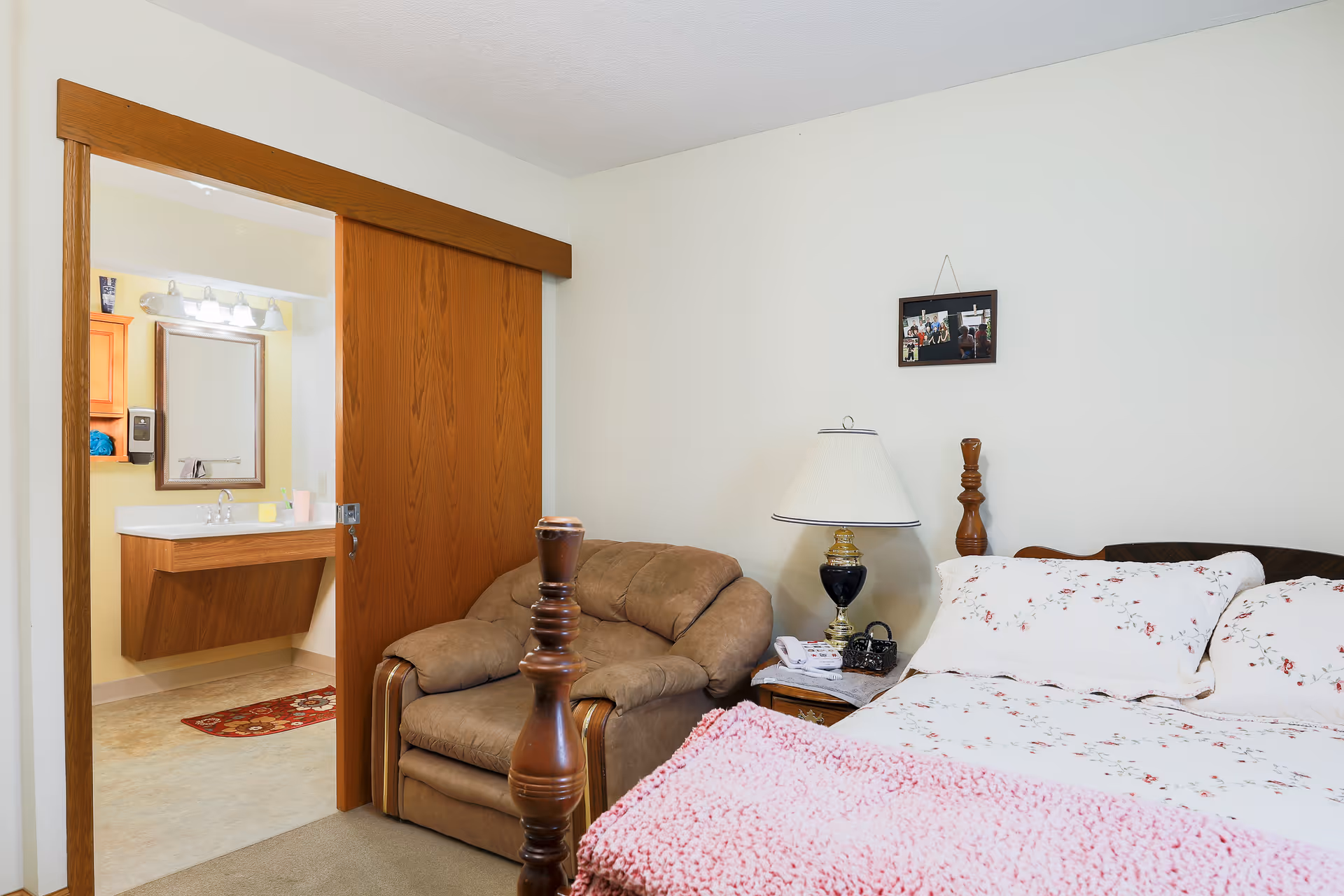 A cozy bedroom in an assisted living facility with a wooden bed frame, floral bedding, a pink blanket, a brown armchair, a nightstand with a lamp and some items, and a sliding wooden door leading to a bathroom with a sink and mirror.