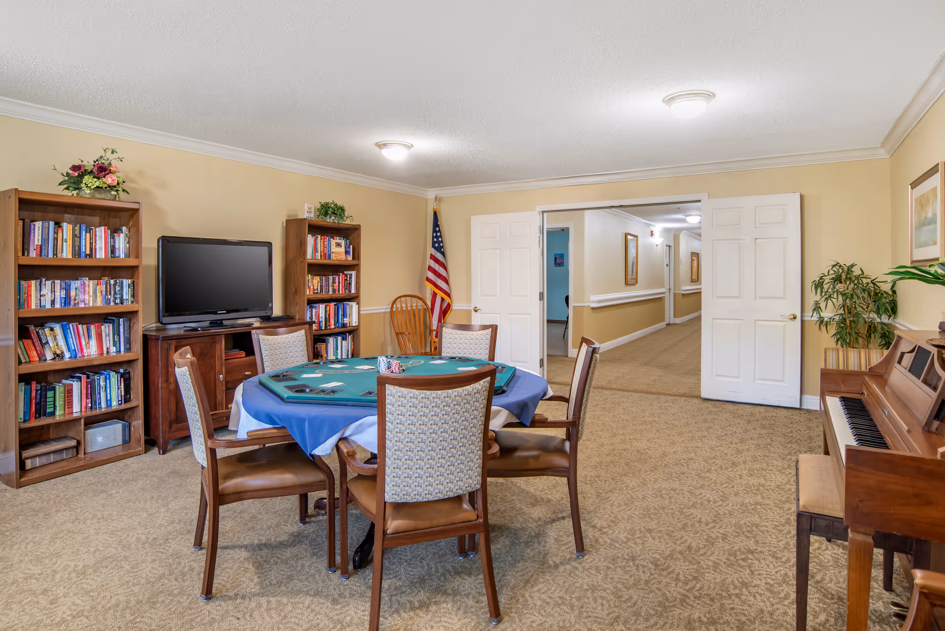 A cozy common room in a senior living facility featuring a round table with a blue tablecloth and a poker game setup, surrounded by six chairs. There are two wooden bookshelves filled with books and a flat-screen TV between them. An American flag stands in the corner near double white doors that open to a hallway. On the right side, there is a wooden piano with a bench and a potted plant beside it. The room has beige walls and carpeted flooring.