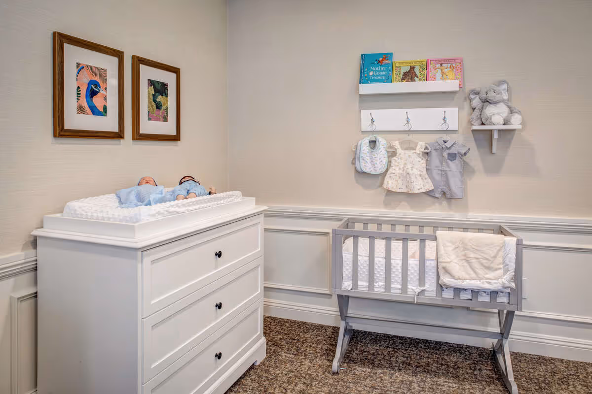 A nursery room with a white changing table holding two baby dolls, a gray crib with a white blanket draped over the side, and a wall with framed bird artwork, hanging baby clothes, children's books, and a stuffed elephant toy on a small shelf.