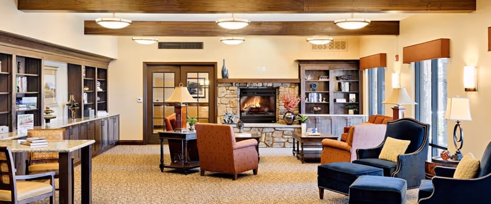 Cozy furnished common room with armchairs, bookshelves, and a stone fireplace in an assisted living facility.
