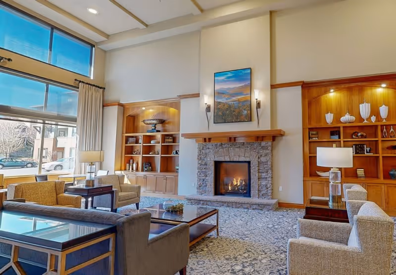A spacious and well-lit living room area with large windows allowing natural light to fill the space. The room features a stone fireplace with a landscape painting above it, flanked by two wall sconces. There are built-in wooden shelves on either side of the fireplace displaying decorative items. Comfortable armchairs and sofas are arranged around a glass coffee table on a patterned carpet. Two table lamps sit on side tables next to the seating.
