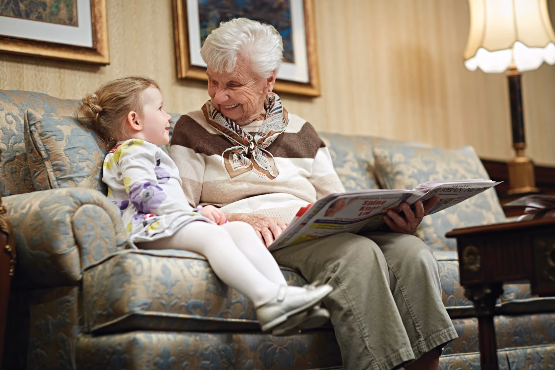 An elderly woman and a young girl sitting together on a patterned sofa in a cozy living room. The woman is holding a newspaper and smiling warmly at the girl, who is looking back at her. The room features framed artwork on the wall and a table lamp with a white shade in the background.