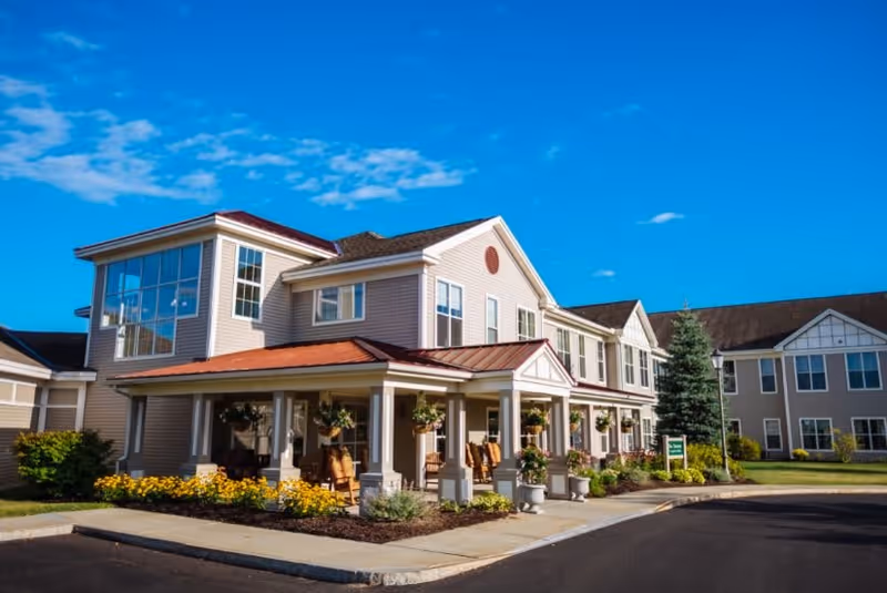 Exterior view of a senior living facility building with beige siding, large windows, and a covered porch with rocking chairs and hanging flower pots. The sky is clear and blue, and there are landscaped flower beds and shrubs around the building.