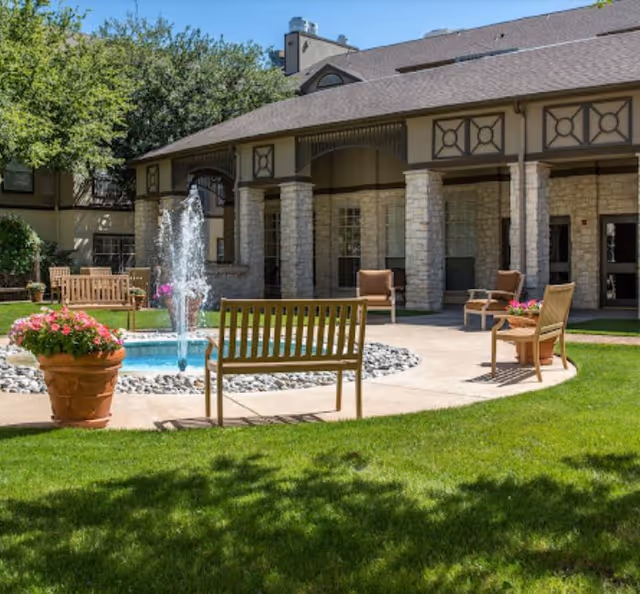 Sunlit courtyard with a central fountain, benches, potted flowers, and the senior living building façade in the background.
