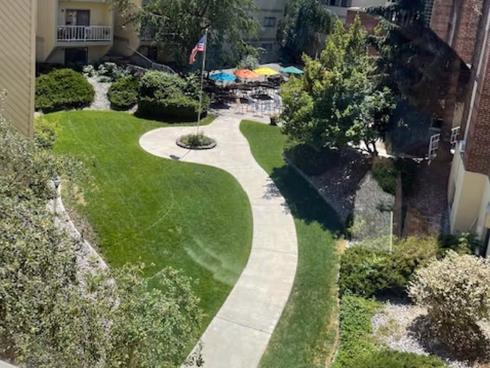 View of a landscaped outdoor garden area with a curved concrete walkway, green grass, bushes, trees, and an American flag on a flagpole. In the background, there are several tables with colorful umbrellas and chairs, surrounded by buildings.