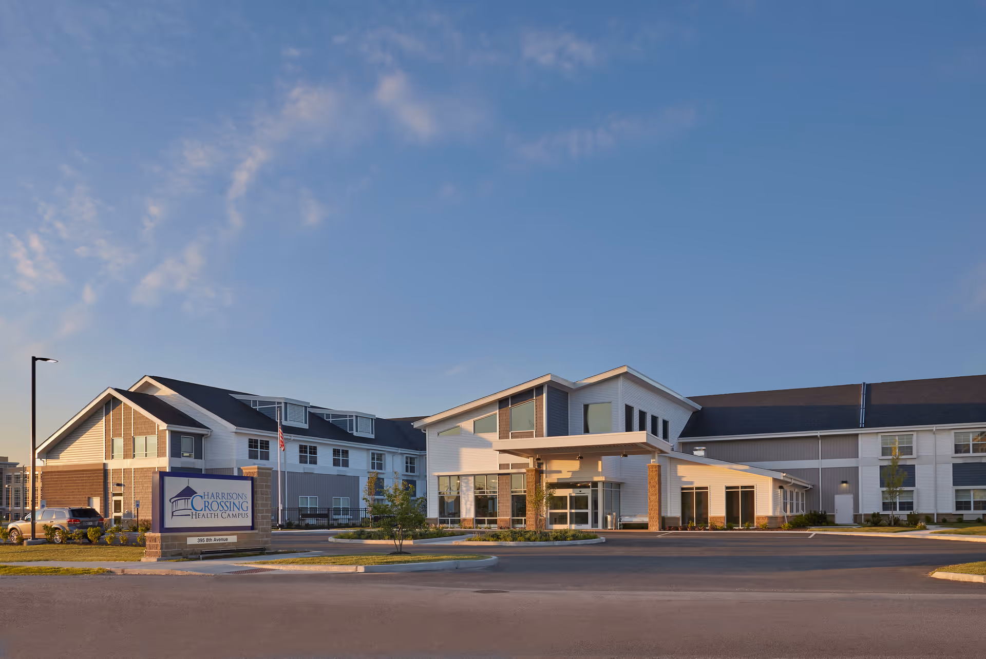 Exterior view of Harrison's Crossing Health Campus building during daylight with clear blue sky. The building has a modern design with large windows and a covered entrance. There is a sign in front displaying the facility's name and address, and a few cars are parked nearby.