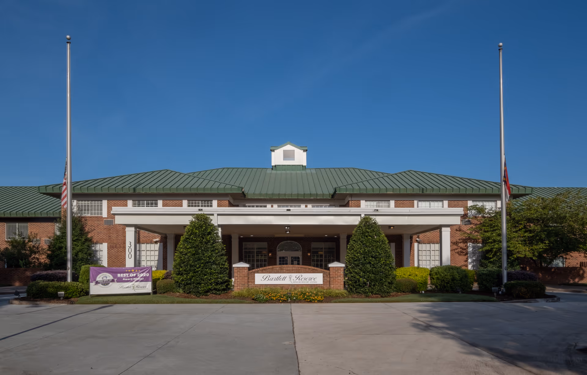 Front exterior view of Bartlett Reserve, a brick building with a green metal roof, two flagpoles with flags at half-mast, and a covered entrance flanked by bushes and landscaping.