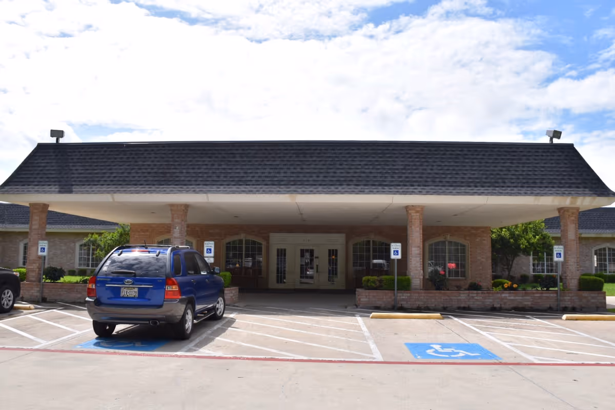Front exterior view of Stonegate Nursing and Rehabilitation facility showing a covered entrance with brick pillars, a blue SUV parked in a handicapped parking spot, and a partly cloudy sky.