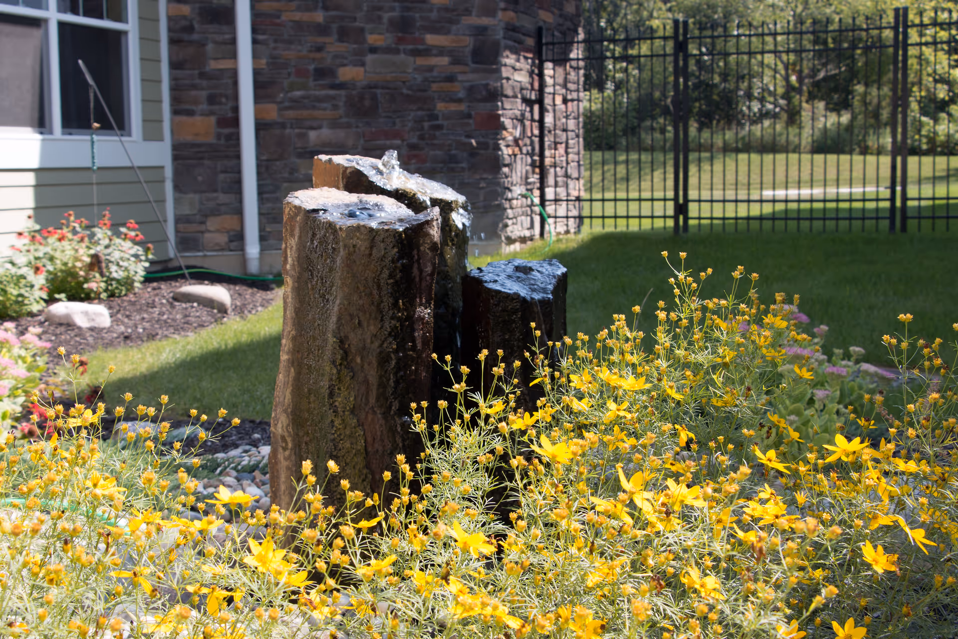 Outdoor garden area with yellow flowers in the foreground, a stone water feature with water flowing on top, green grass, and a building with stone and siding walls in the background, along with a black metal fence.