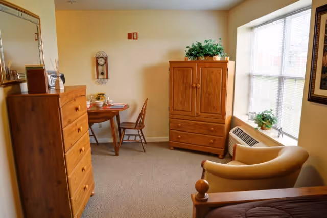 Sunlit furnished senior living bedroom with wooden dresser, armoire, small table and armchair by a window.