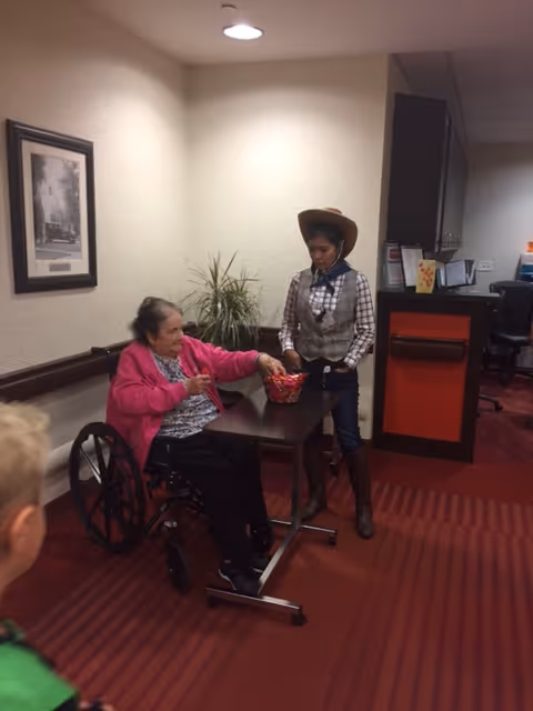 An elderly woman in a wheelchair wearing a pink jacket is sitting at a small table reaching into a basket held by a young woman dressed in a cowboy hat, plaid shirt, vest, and boots. They are in an indoor setting with red carpet and beige walls, with a framed picture and a plant in the background.