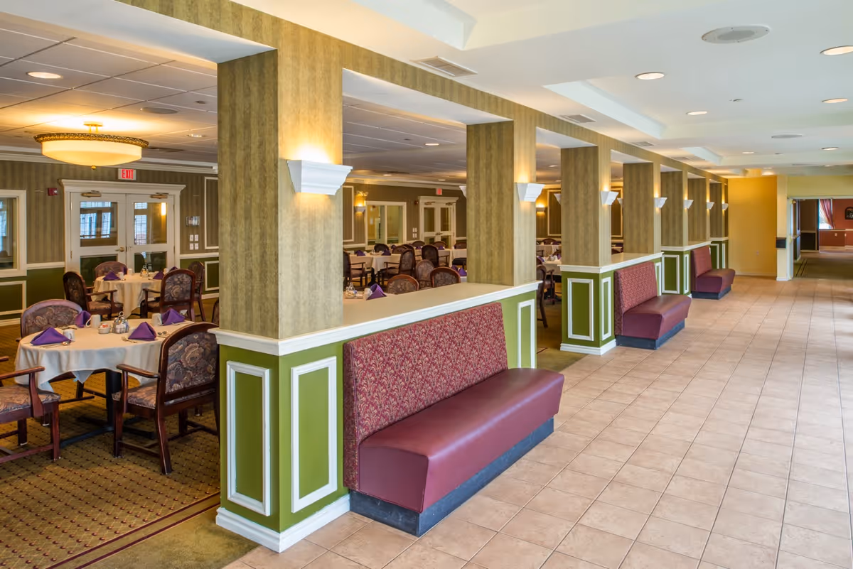 Spacious assisted living dining room with round tables set for meals and upholstered banquette seating along a tiled walkway.