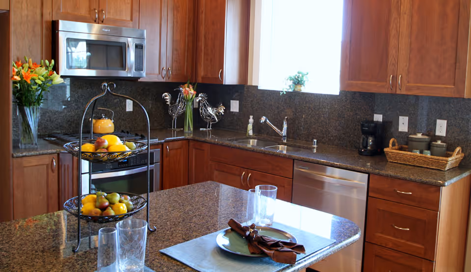 A modern kitchen with wooden cabinets, granite countertops, and stainless steel appliances including a microwave, oven, and dishwasher. The kitchen island has a two-tiered fruit basket with assorted fruits, two clear drinking glasses, and a place setting with a plate and brown napkin. There are flowers in vases on the counter and a coffee maker with a basket of mugs on the right side.