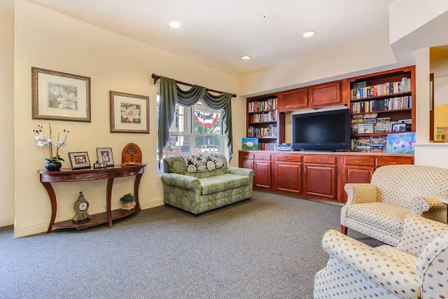 A cozy living room area in an assisted living facility featuring a green upholstered loveseat, two beige armchairs with patterned fabric, a wooden console table with framed photos and decorative items, and a built-in wooden entertainment center with a flat-screen TV and shelves filled with books and games. A window with green curtains lets in natural light.