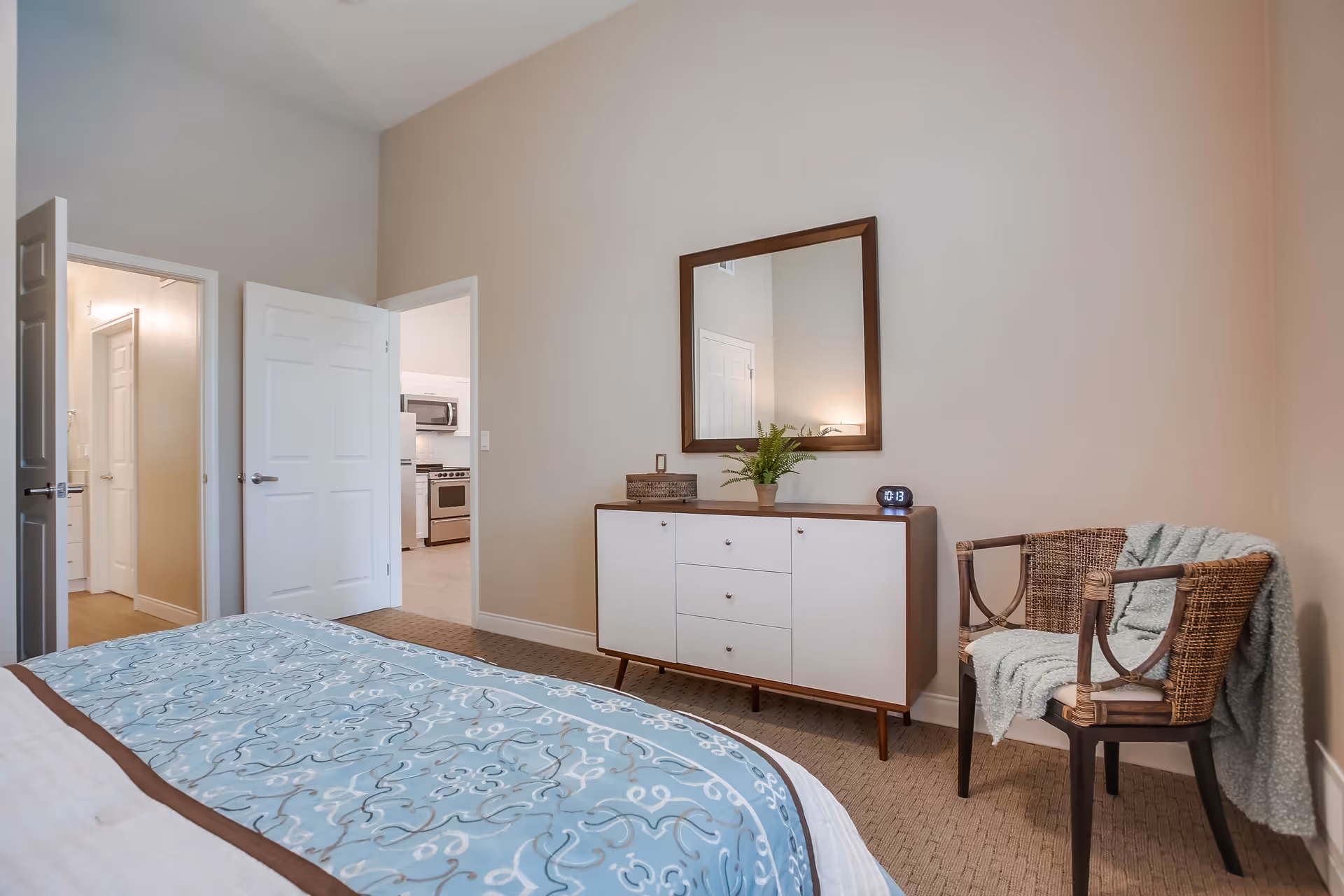 A bedroom with a bed covered in a blue patterned blanket, a white dresser with a mirror above it, a small plant and a digital clock on the dresser, and a wicker chair with a light blue throw blanket draped over it. Two open doors reveal a hallway and a kitchen area.