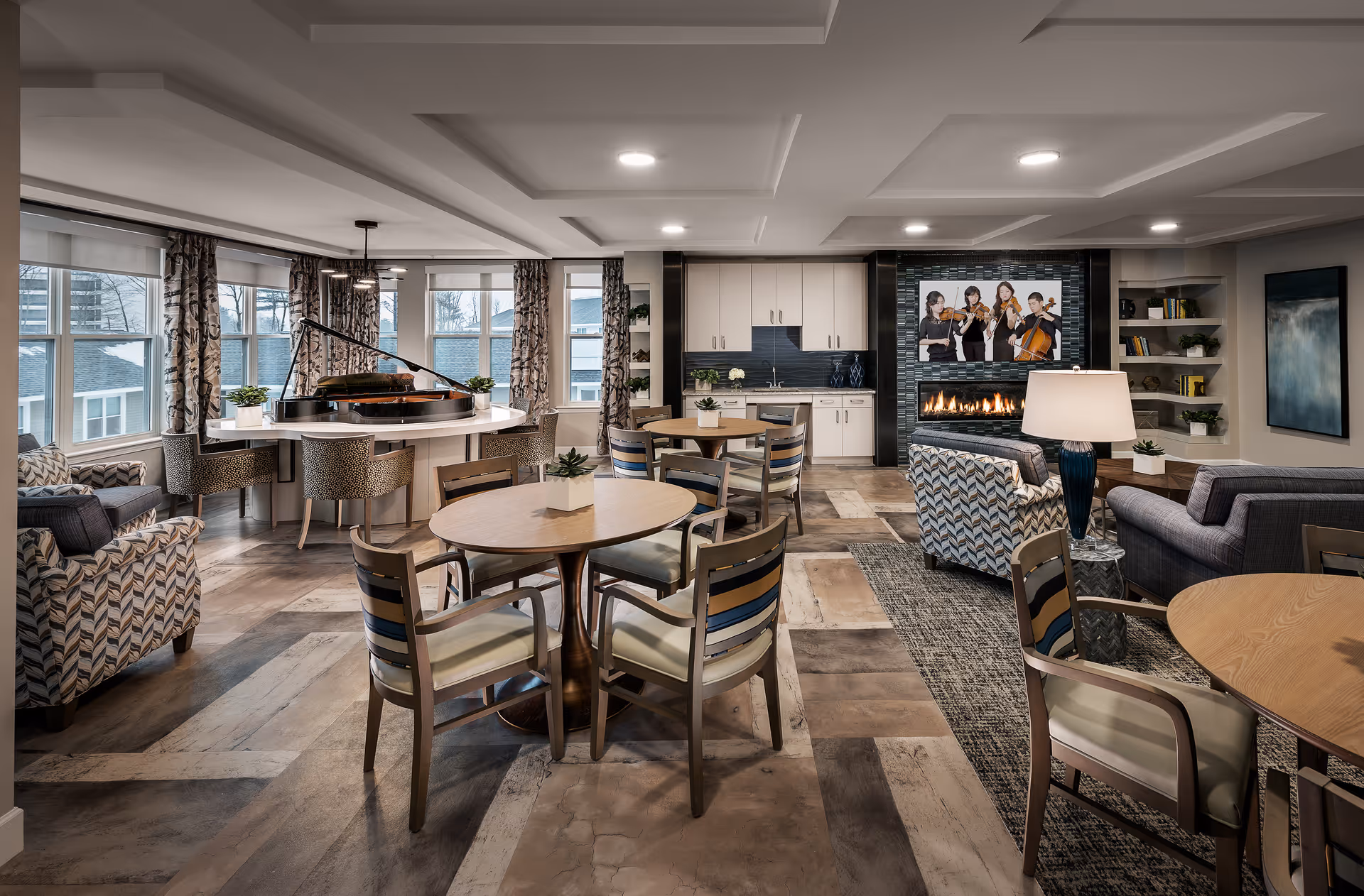 A spacious and well-lit senior living common area featuring multiple round wooden tables with chairs, comfortable patterned armchairs, a grand piano near large windows with curtains, a modern fireplace with a TV above it displaying a string quartet, built-in shelves with decorative items, and a small kitchenette with white cabinets in the background.