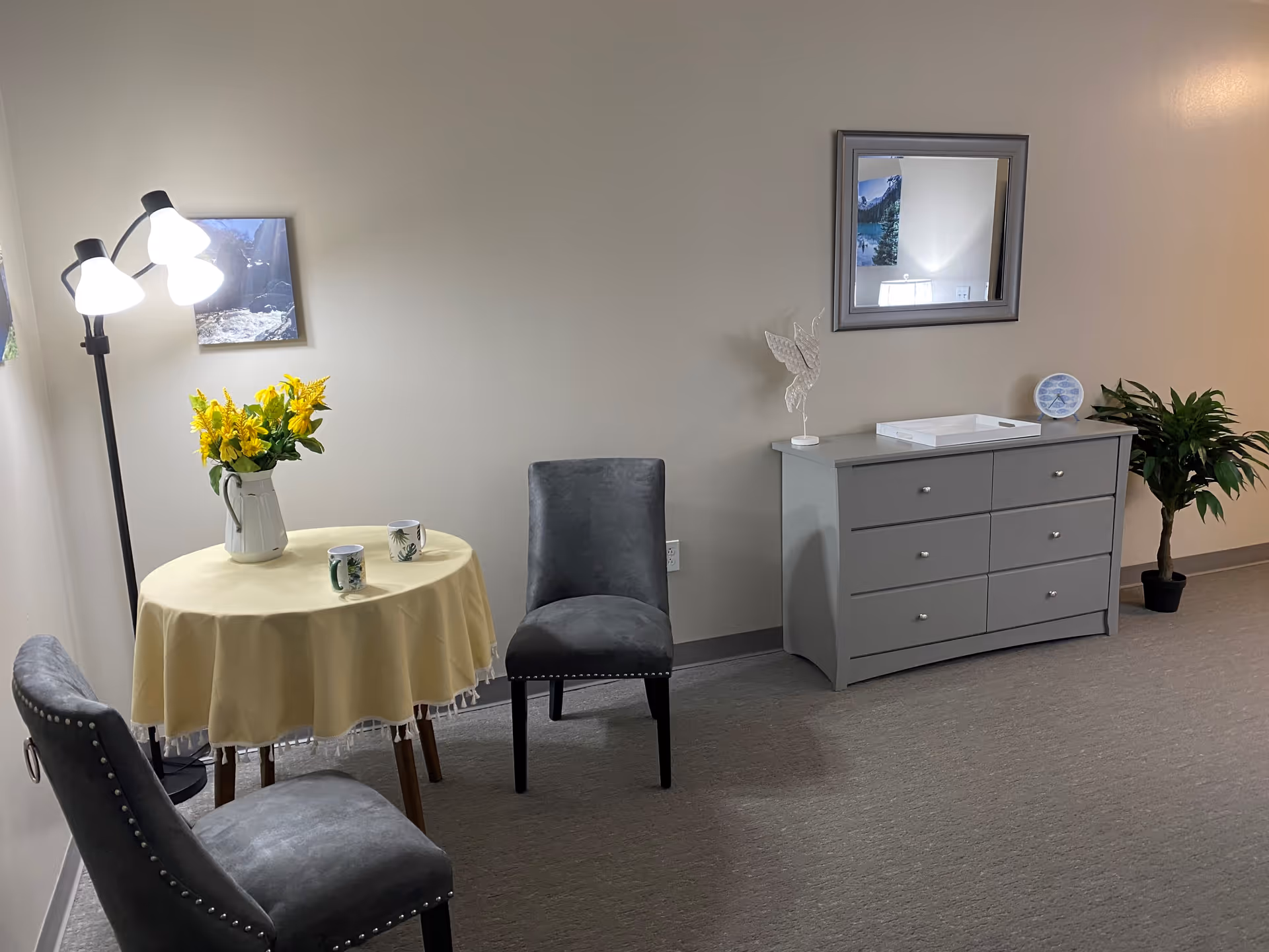 A small sitting area in a room with two gray upholstered chairs around a round table covered with a yellow tablecloth. On the table is a white pitcher with yellow flowers and two coffee mugs. To the right is a gray dresser with six drawers, a white tray, a decorative bird sculpture, and a small plate. Above the dresser is a rectangular mirror. A potted plant is placed on the floor next to the dresser. A floor lamp with three lights stands near the table, and a framed picture hangs on the wall behind it.