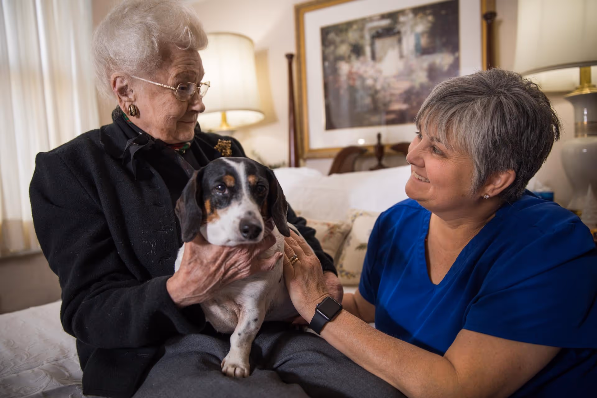 An elderly woman sitting on a bed holding a small dog while a caregiver in blue scrubs smiles and gently pets the dog in a warmly lit bedroom setting.