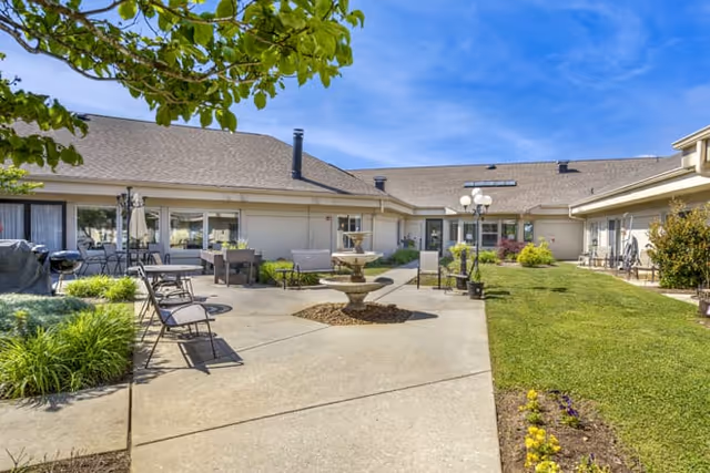 Sunny courtyard at a senior living facility featuring a central fountain, patio seating, and surrounding single-story building.