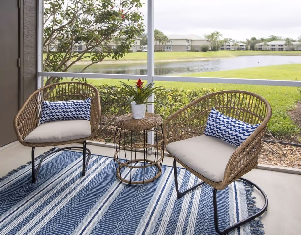 A screened-in patio area with two wicker chairs featuring beige cushions and blue patterned pillows, a small round wicker table with a potted plant on top, and a blue and white striped rug. Outside the patio, there is a grassy area with a pond and residential buildings in the background.