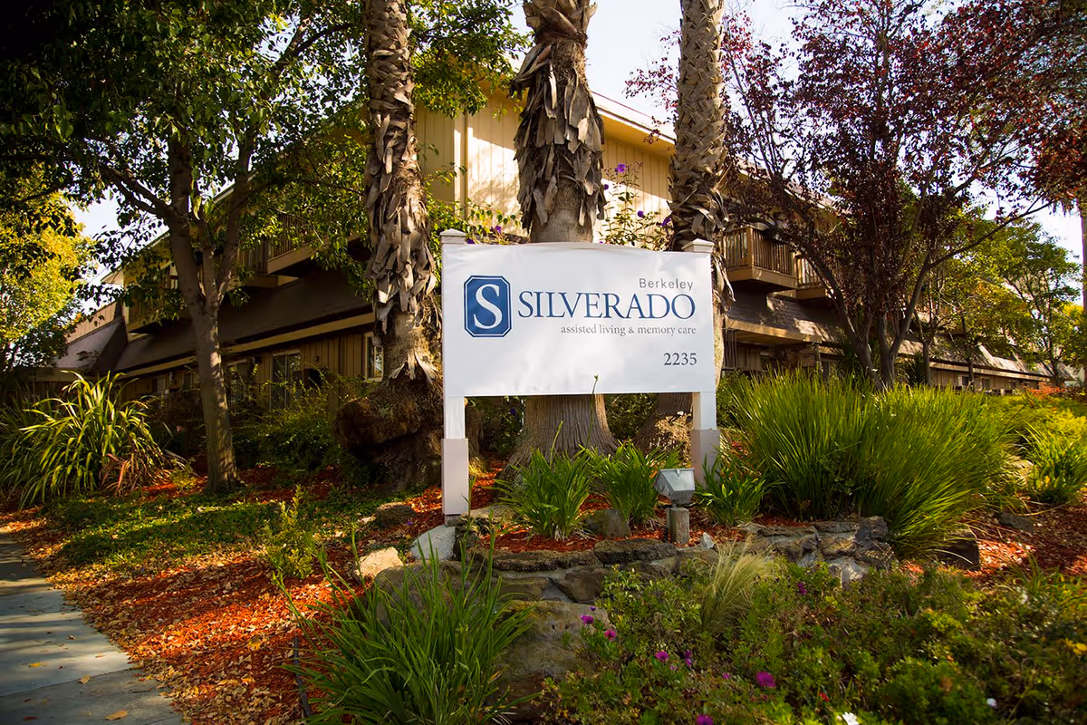 Outdoor view of Silverado Berkeley Memory Care Community sign surrounded by trees, plants, and landscaping with a building partially visible in the background.